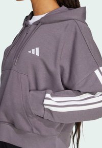 Fioletowa bluza z kapturem, z przednią kieszenią, sznurkiem na kapturze oraz białymi paskami na rękawach. Posiada białe logo na klatce piersiowej. Wykonana z miękkiej mieszanki bawełny.