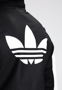Černá bunda s velkým bílým grafickým prvkem loga Adidas trefoil a třemi vodorovnými pruhy pod designem. Hladká textura.