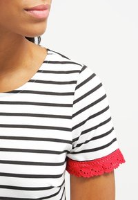 Haut à manches courtes rayé noir et blanc avec une bordure en dentelle rouge aux manches. Matière en coton douce, encolure ronde et design ajusté.