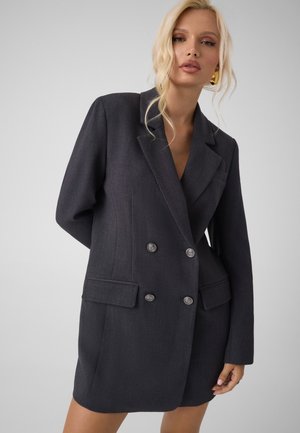 Femme aux cheveux blonds portant une robe blazer oversize à double boutonnage gris foncé avec des boutons argentés sur un fond clair uni.