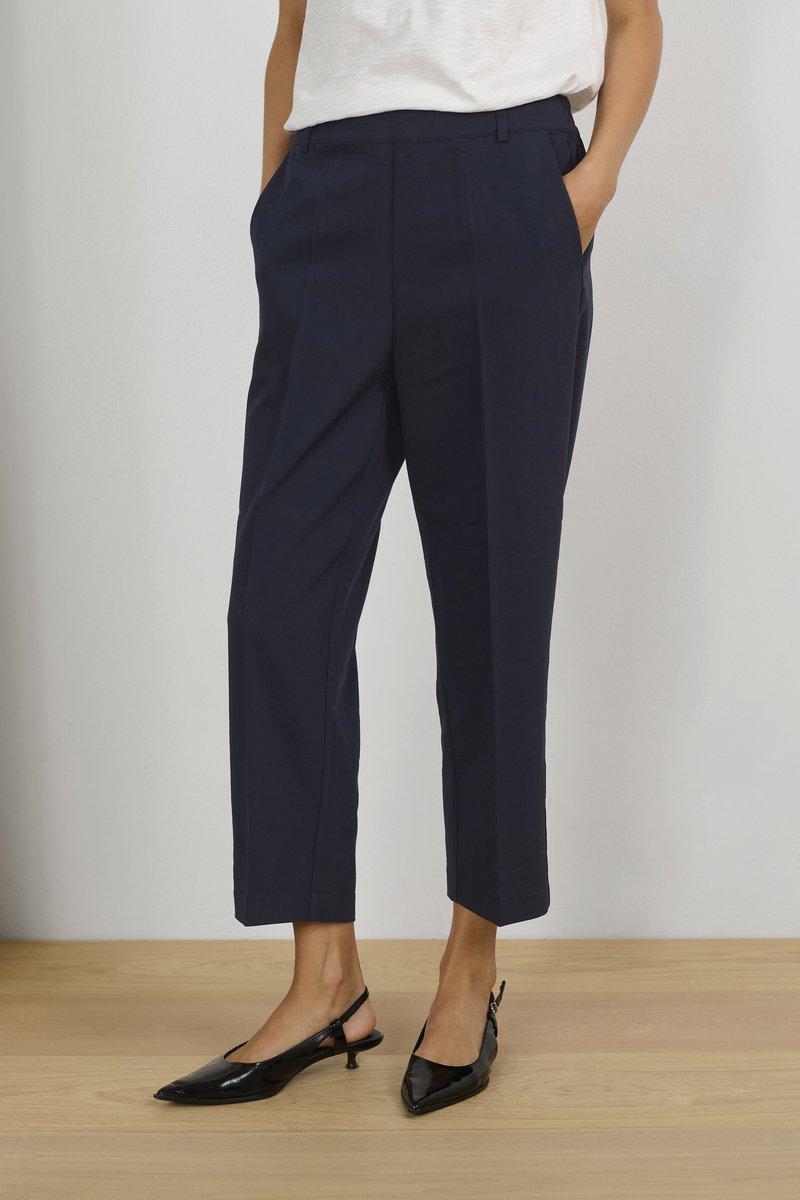 Marineblauwe cropped broeken met voorplooien en zijzakken, gecombineerd met zwarte gepoedercoat slingback schoenen met spitse neuzen. Vlakke houten vloer.