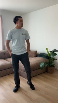 Bărbat cu mâinile în buzunare stă pe podea de lemn lângă o canapea maro și o plantă verde cu frunze într-o cameră luminoasă, decorată minimalist.