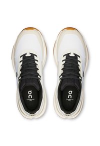 Baskets de sport blanches et grises avec des lacets noirs, un dessus texturé et des semelles en caoutchouc marron. Présente un logo sur la semelle intérieure et un design épuré.