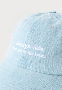 Ανοιχτό μπλε τζιν καπέλο με λευκή κεντημένη επιγραφή "πάντα αργώ αλλά αξίζει η αναμονή" στην μπροστινή πλευρά.