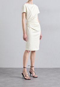 Knee-length, krämfärgad klänning med rund halsringning, korta ärmar och samlade sidodetaljer, i kombination med svarta remsandaler med hög klack.