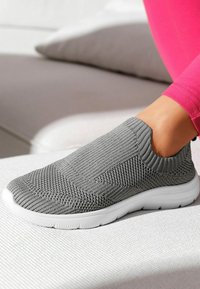 Graue Slip-On-Sneaker mit strukturiertem Strick-Obermaterial, elastischem Kragen und gepolsteter, weiße Sohle, auf einer hellen Oberfläche platziert.