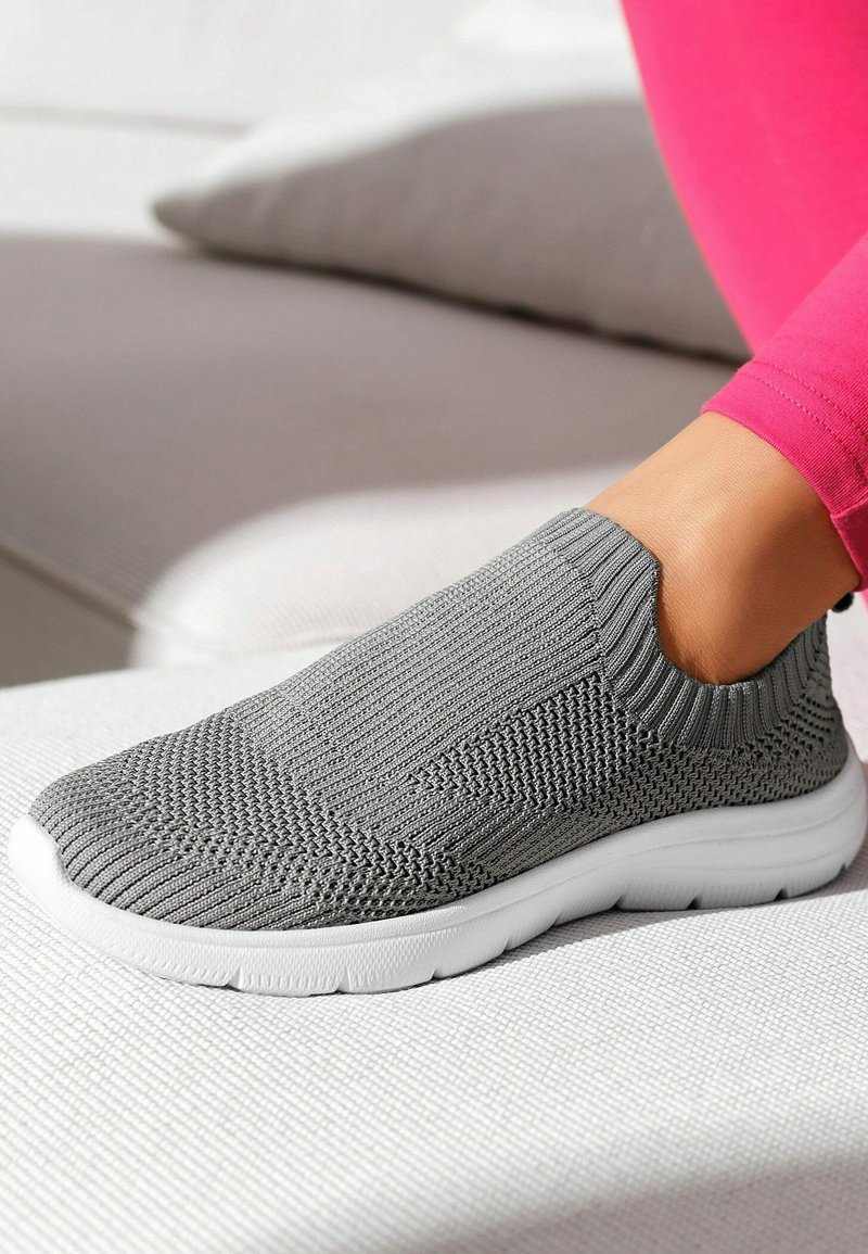 Graue Slip-On-Sneaker mit strukturiertem Strick-Obermaterial, elastischem Kragen und gepolsteter, weiße Sohle, auf einer hellen Oberfläche platziert.