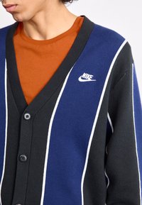 Sininen ja musta neuletakki valkoisilla raidoilla, jossa on V-aukko, viisi nappia ja pieni valkoinen Nike-logo rinnassa.