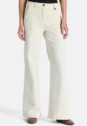 Femme portant un pantalon large en velours côtelé crème taille haute avec poches avant et des chaussures marron en peau de serpent, debout devant un fond uni.