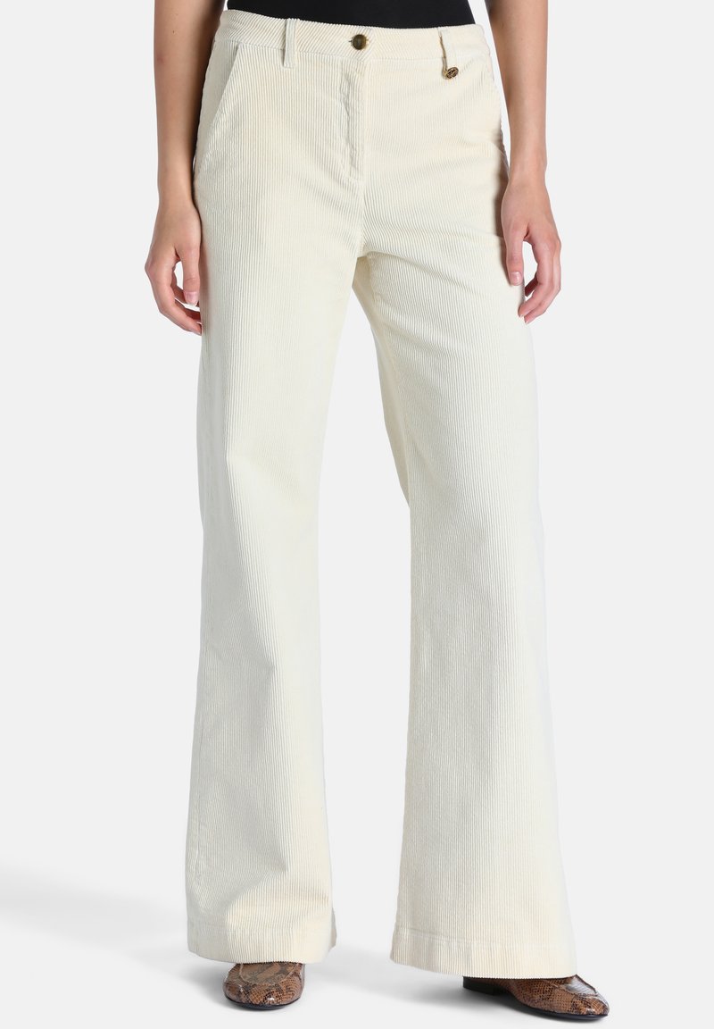 Femme portant un pantalon large en velours côtelé crème taille haute avec poches avant et des chaussures marron en peau de serpent, debout devant un fond uni.