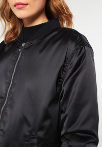 Svart bomberjacka med en glänsande satinfinish, ribbad krage och dragkedjestängning. Har sidofickor och detaljerad sömnadsaccent.