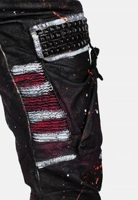 Zwarte versleten broeken met rode en zilveren strepen, textuurelementen en een gestifte zakdetail. Bevat verfspatten en heeft een losse pasvorm.