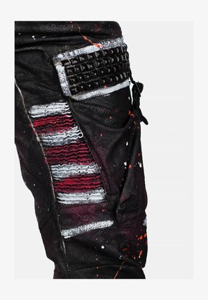 Pantaloni neri invecchiati con strisce rosse e argento, dettagli testurizzati e una tasca con borchie. Presentano macchie di vernice e una vestibilità rilassata.