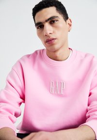 Růžová mikina s vroubkovaným kulatým výstřihem, která má na hrudi vyvýšené bílé logo "GAP". Hladká látka, volný střih a dlouhé rukávy.