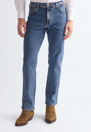 Mann trägt gerade geschnittene blaue Jeans und braune Wildleder-Stiefeletten, steht vor einem einfarbigen hellen Hintergrund.