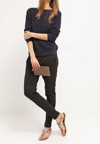 Pull à côtes marine, pantalon noir fuselé et sandales argentées à brides. Tient une pochette marron à texture lisse.