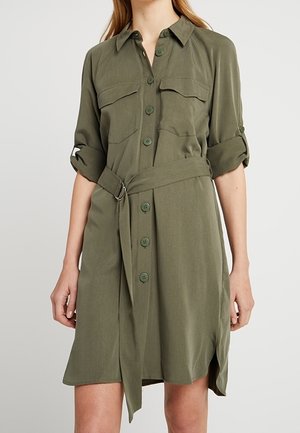 Femme portant une robe boutonnée olive longueur genou, avec des manches retroussées et une ceinture en tissu assortie à la taille.