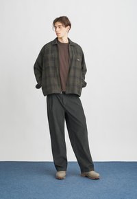 Mlad moški stoji na modrem preprogo, oblečen v temno karirasto jakno z zadrgo, rjav srajco, ohlapne črne hlače in bež čevlje ob beli steni.