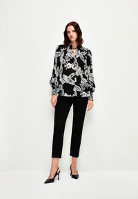 Schwarze Bluse mit weißem Paisleymuster, langen Ärmeln, Bindeband am Ausschnitt, kombiniert mit schwarzen Hosen und schwarzen High-Heels. Glatte Textur.