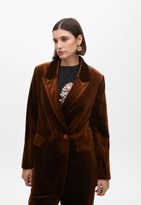 Brun sammetsp blazer med dubbelknäppt design, slagkrage och två framfickor. Tyget har en jämn och rik textur.