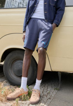 Grijze sportbroek met turquoise accenten, gecombineerd met beige sneakers en witte sokken, tegen een achtergrond van een vintage geel voertuig.