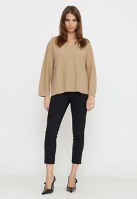 Brauner Strickpullover mit V-Ausschnitt, kombiniert mit engen schwarzen Hosen und spitzen schwarzen High Heels, was einen lässigen, aber gepflegten Look erzeugt.
