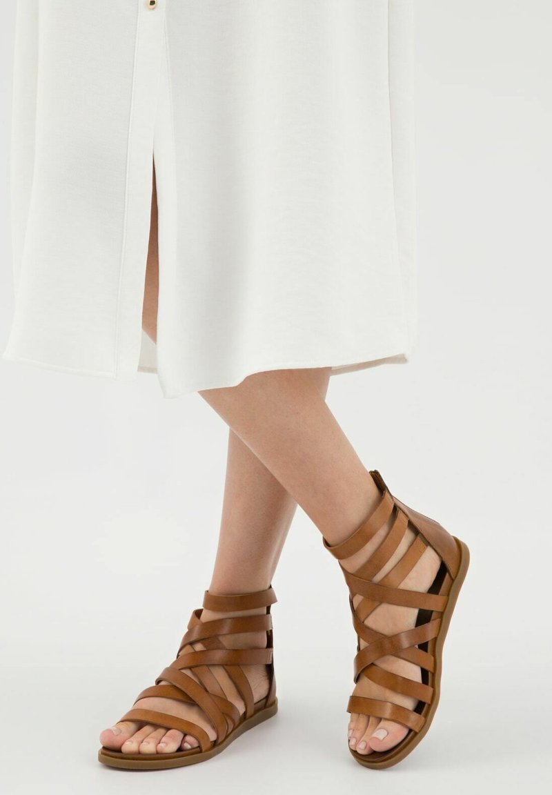 Sandali strappy in pelle marrone con più cinghie, punta aperta, suola piatta e altezza alla caviglia, progettati per il comfort e la traspirabilità.