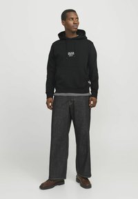 Schwarzer Hoodie mit Logo, graue Innenschicht und weite dunkelblaue Jeans. Braune Schuhe mit strukturierter Sohle, stehend vor einem neutralen Hintergrund.