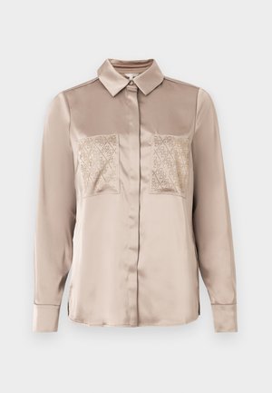 Beige satijnen blouse met lange mouwen, een puntige kraag, verborgen knopen en twee voorzakken met een subtiel diamanten patroon.