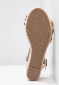 Vue de la semelle d'une sandale compensée beige avec semelle extérieure en caoutchouc texturé et bride cheville, posée verticalement sur un fond blanc.