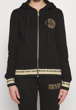 Personne portant un survêtement noir Versace Jeans Couture avec détails de logo dorés, fermeture éclair dorée et finitions dorées de marque aux poignets, à l'ourlet et au pantalon.