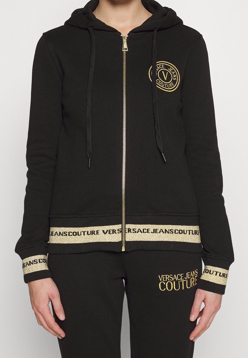 Personne portant un survêtement noir Versace Jeans Couture avec détails de logo dorés, fermeture éclair dorée et finitions dorées de marque aux poignets, à l'ourlet et au pantalon.