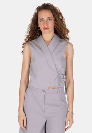 Donna con capelli ricci che indossa un gilet incrociato grigio chiaro senza maniche e pantaloni su misura abbinati su uno sfondo semplice.