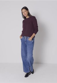 Burgundy tröja, lösa blå denimjeans och svarta ankelstövlar. Modellen har långt mörkt hår. Neutral bakgrund.