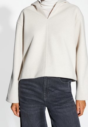 Persona con un jersey con capucha color crema de manga larga y vaqueros oscuros de talle alto, mostrado desde los hombros hasta las caderas.