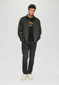 Black pinstriped shirt, black t-shirt with gold text, dark jeans, and beige sneakers, creating a casual layered outfit with contrasting elements.