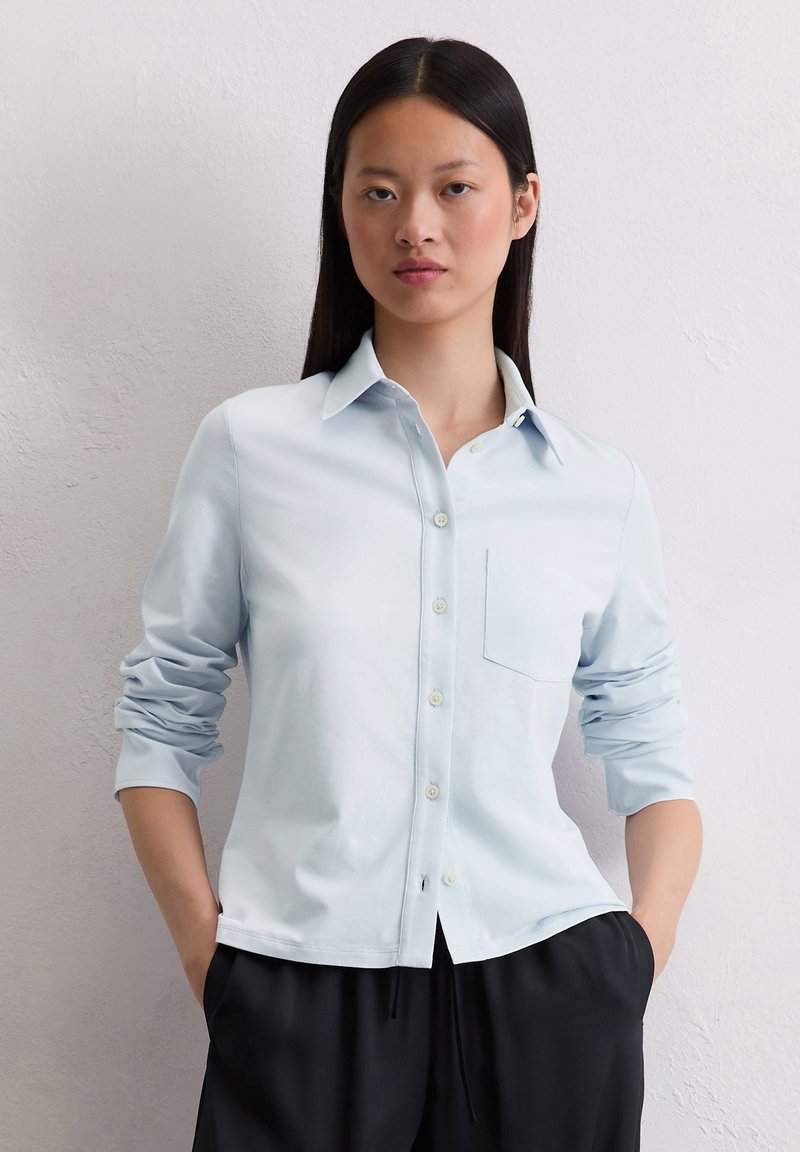 Camicia azzurro chiaro, con bottoni, realizzata in un tessuto morbido, con maniche lunghe arrotolate e taschino sul petto. Presenta un colletto classico e una leggera lucentezza.