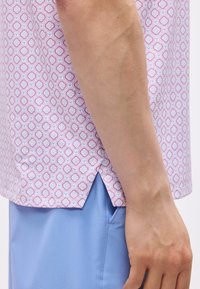 Ανδρικός βραχίονας δίπλα σε ανοιχτό ροζ πουκάμισο με μοτίβο και πλαϊνή σχισμή, και ανοιχτό μπλε παντελόνι με εμφανές ραφή τσέπης.