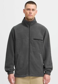 Veste en polaire grise avec col montant, fermeture zipp�ée à l'avant et deux poches latérales. Comprend une poche poitrine zippée et des poignets élastiqués.