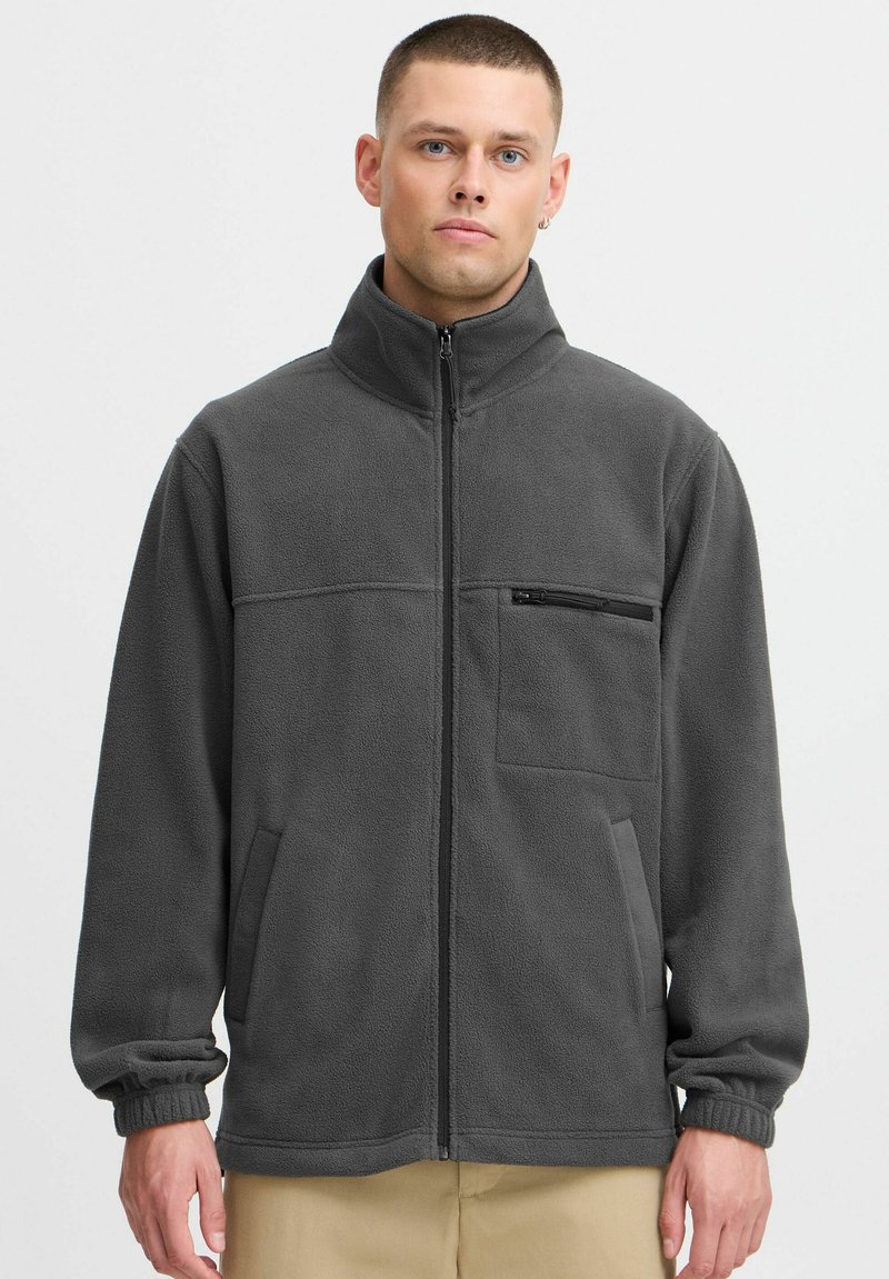 Veste en polaire grise avec col montant, fermeture zippée à l'avant et deux poches latérales. Comprend une poche poitrine zippée et des poignets élastiqués.