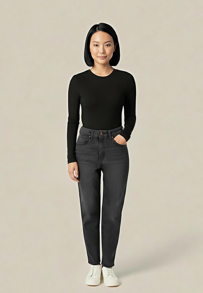 Kobieta z czarnym bobem, ubrana w czarną bluzkę z długim rękawem, ciemnoszare d�żinsy z wysokim stanem i białe buty, stojąca na tle jednolitego beżowego tła.