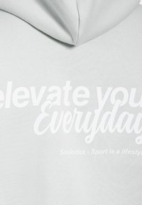 Jasnoszara bluza z kapturem z dużym białym napisem: "Elevate your Everyday" oraz "Smilodox - Sport to styl życia." Miękka, gładka tekstura materiału.