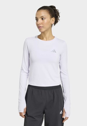 Frau mit lockigem Haar im Pferdeschwanz, die ein weißes langärmeliges Sportoberteil und schwarze Hosen mit elastischem Bund trägt und nach links schaut.