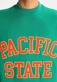 Grön tröja med texten "PACIFIC STATE" i orange och gult, bärs av en person och är beskuren under halsen.