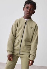 Olea rohelise lukuga sviiter, millel on kaks esikotti, rippuv varrukate ots ja rinna peal logo "BAKER by TED BAKER". Sile tekstuur.