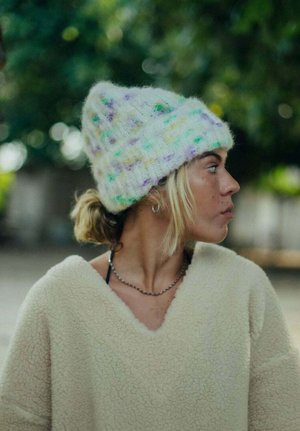 Młoda osoba z jasnymi włosami, w kolorowej dzianinowej czapce, kremowym polarze i naszyjniku z łańcuszka, patrzy w prawo na zewnątrz na tle rozmytej zieleni.