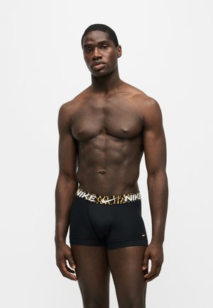 Μυώδης άνδρας που φοράει μαύρα boxer briefs Nike με ζώνη με ζωικό μοτίβο, στέκεται μπροστά σε απλό ανοιχτό φόντο.