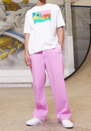Persoon in een loszittend wit T-shirt met een kleurrijk design en brede roze broek staand bij een skatehelling binnen.