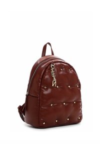 Petit sac à dos en cuir marron avec des clous en forme de cœur dorés et un charm en forme de lettre dorée suspendu à la fermeture éclair avant.