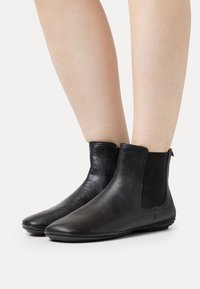 Botas de cuero negro hasta el tobillo con paneles laterales elásticos, textura suave, punta redonda y suela de perfil bajo. Diseño minimalista con un acabado elegante.